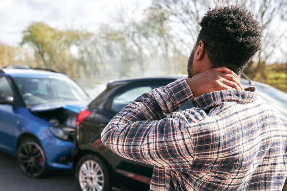 An injured person stands next to a damaged car, holding their neck in pain after an accident