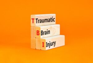 Symbolizing traumatic brain injury (TBI), wooden blocks with the concept words "TBI traumatic brain injury" are arranged on a vibrant orange table.