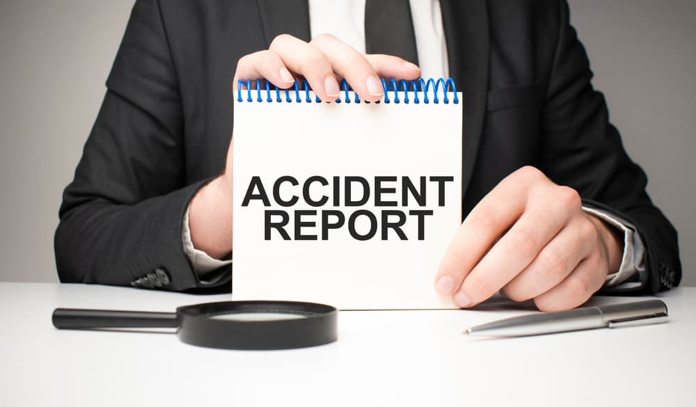 A lawyer writes in a notebook with a silver pen, holding a card labeled "Accident Report." The lawyer is gathering necessary evidence.