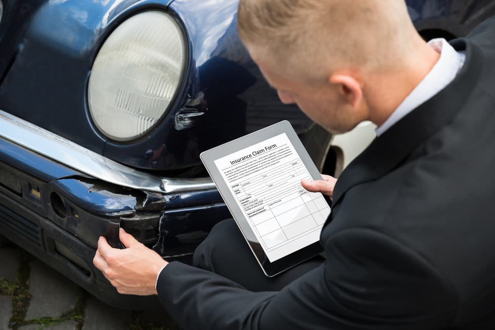 Insurance agent inspecting a damaged car while using a digital tablet to complete an insurance claim form.