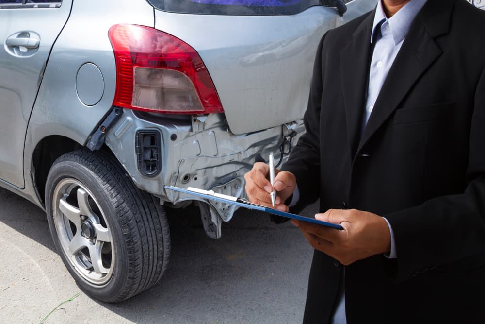 A side view of an insurance officer writing on a clipboard while an insurance agent examines a black car after an accident.