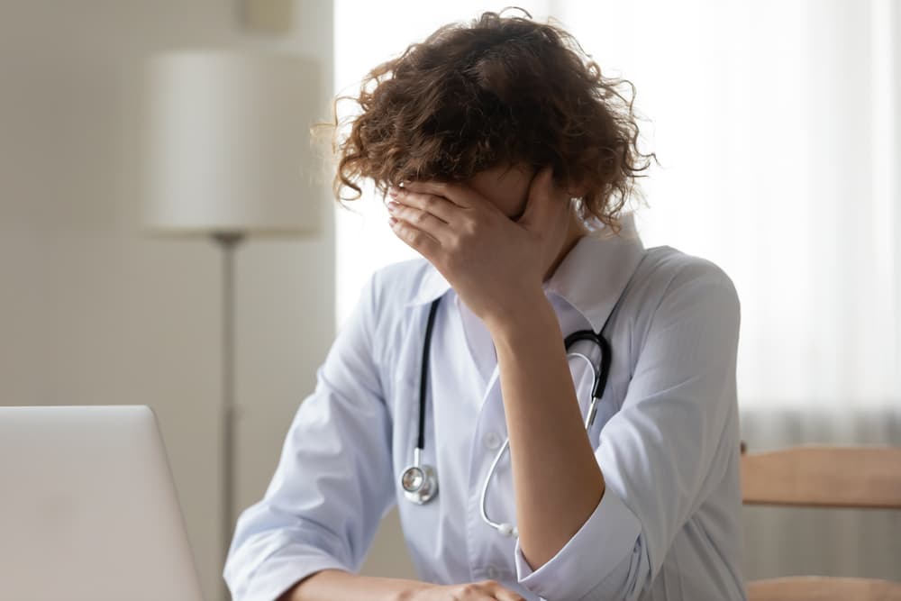 A stressed female doctor with her hand covering her face, sitting at a desk with a laptop.