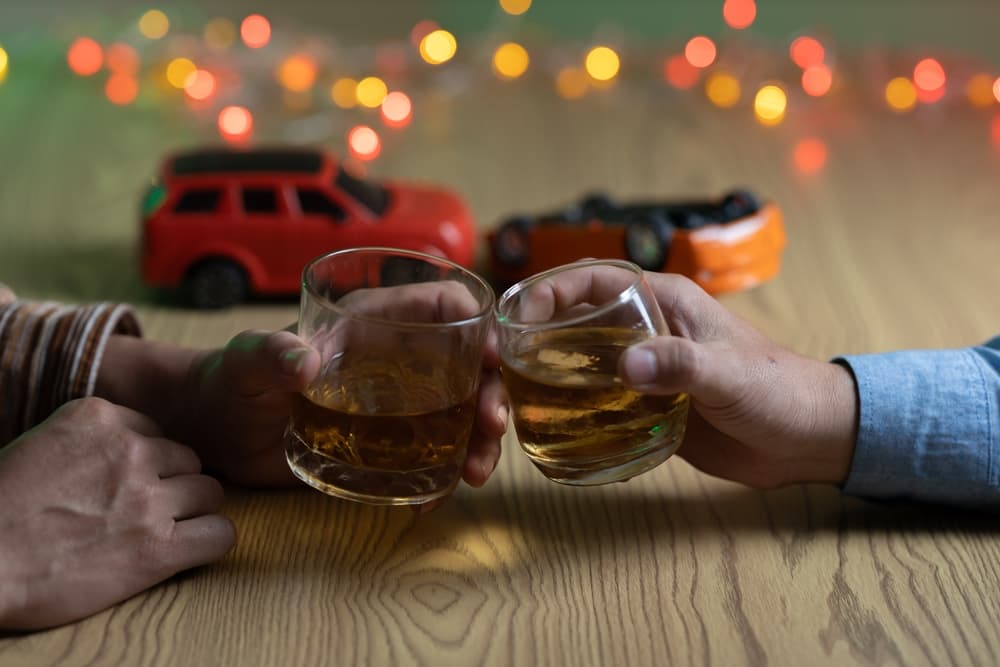 Two men clink whiskey glasses while in front of a bar in a restaurant. With a small car model on the table.