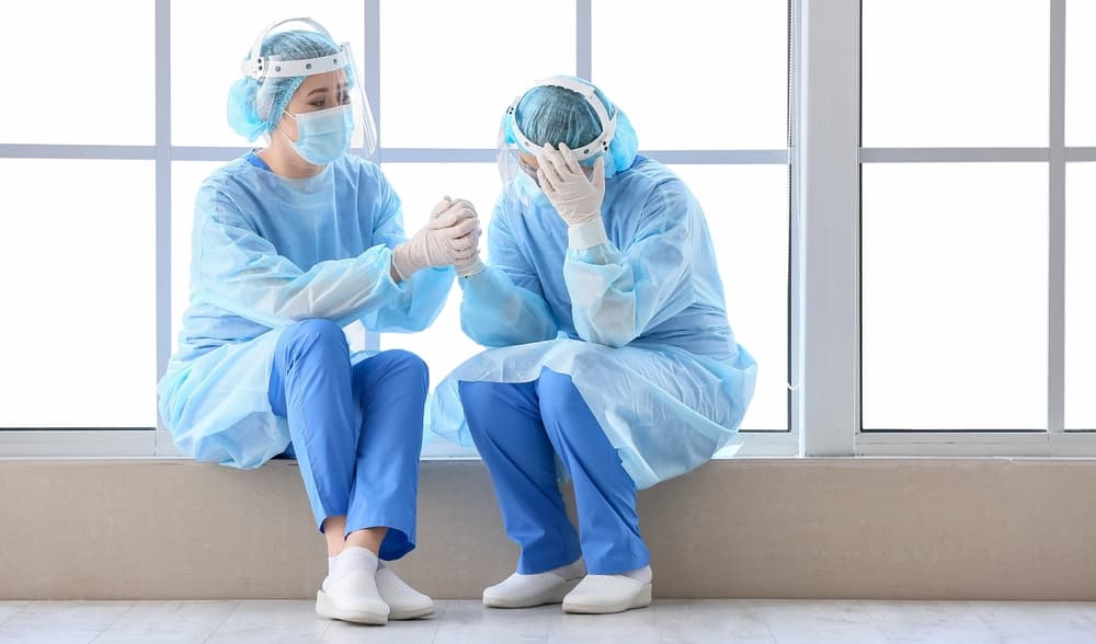 Two medical professionals in protective gear, one comforting the other, sitting by a large window.