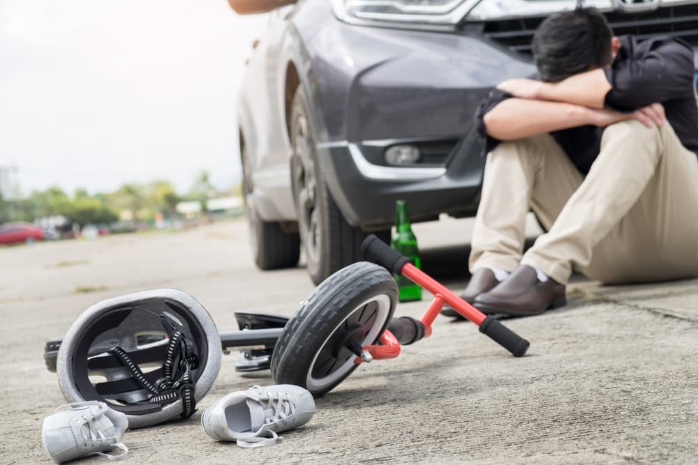 A man sitting by a car, looking distressed, with a tricycle and helmet on the ground.