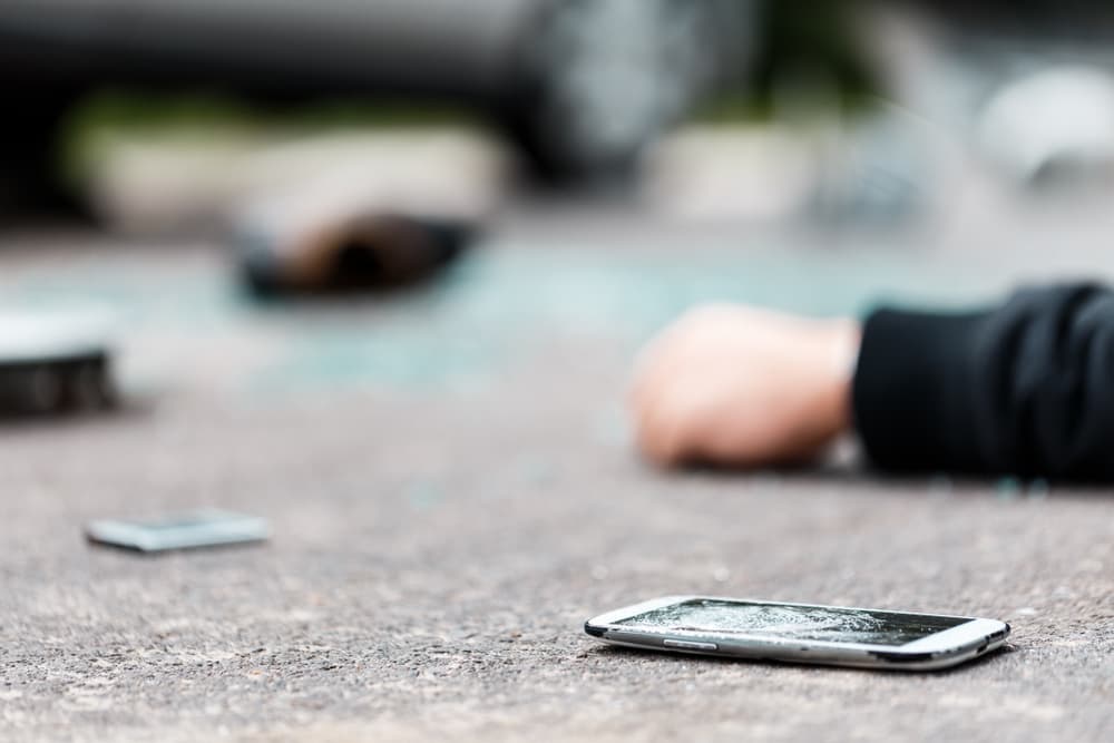 A broken smartphone lying on the ground near a person's outstretched hand, depicting an accident scene.