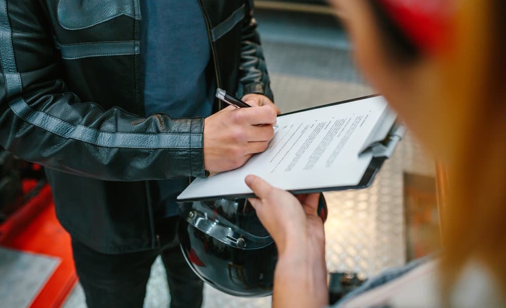 A motorcyclist signing legal documents on a clipboard, possibly related to a wrongful death claim.